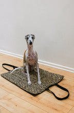 A whippet dog sits on a leopard-print dog mat with black handles, placed on a light wood floor against a plain wall.