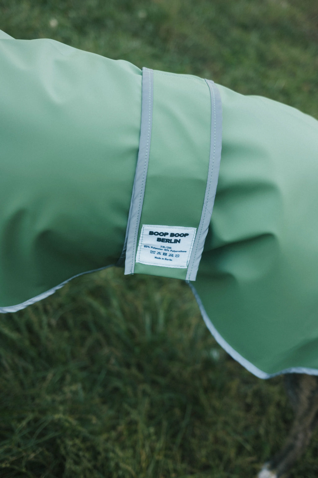 A close-up of a sage green dog raincoat with a white and gray adjustable strap, featuring a white 'BOOP BOOP DESIGN' brand label, set against a blurred green grass background.