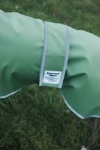 A close-up of a sage green dog raincoat with a white and gray adjustable strap, featuring a white 'BOOP BOOP DESIGN' brand label, set against a blurred green grass background.