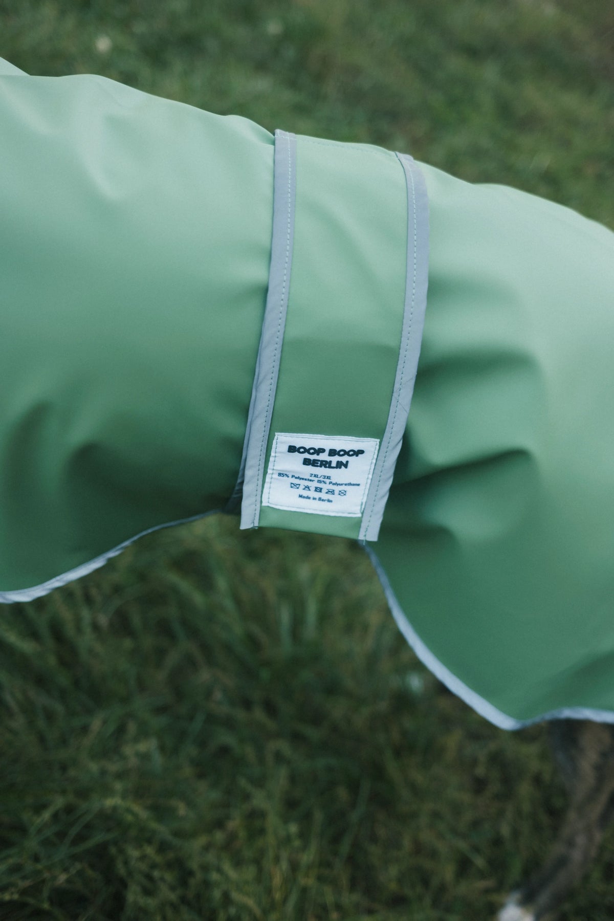 A close-up of a sage green dog raincoat with a white and gray adjustable strap, featuring a white 'BOOP BOOP DESIGN' brand label, set against a blurred green grass background.
